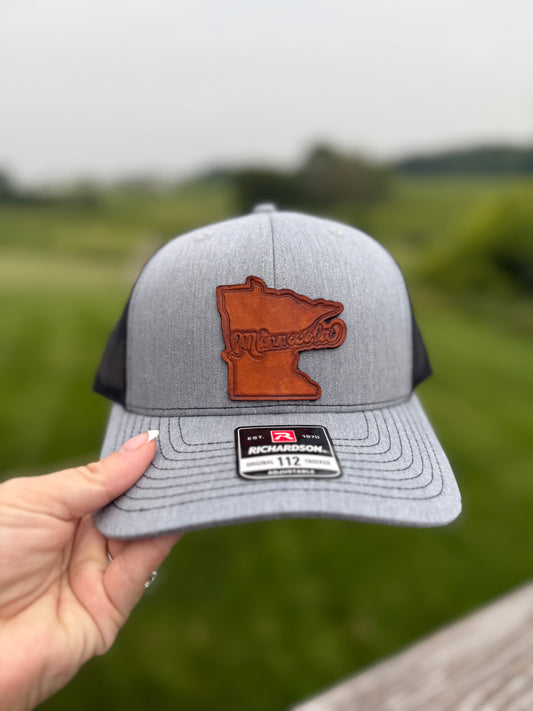 Gray and black cap with a brown Minnesota-shaped patch held by a hand against a blurred green background.