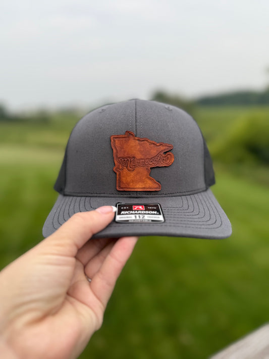 Gray cap with a brown leather Minnesota shape patch held in front of a blurred green field.
