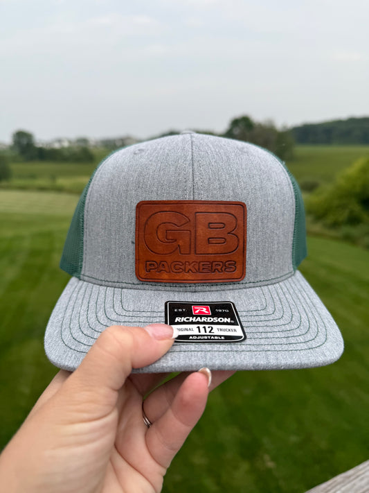 Gray and green cap with leather patch held in front of a grassy field