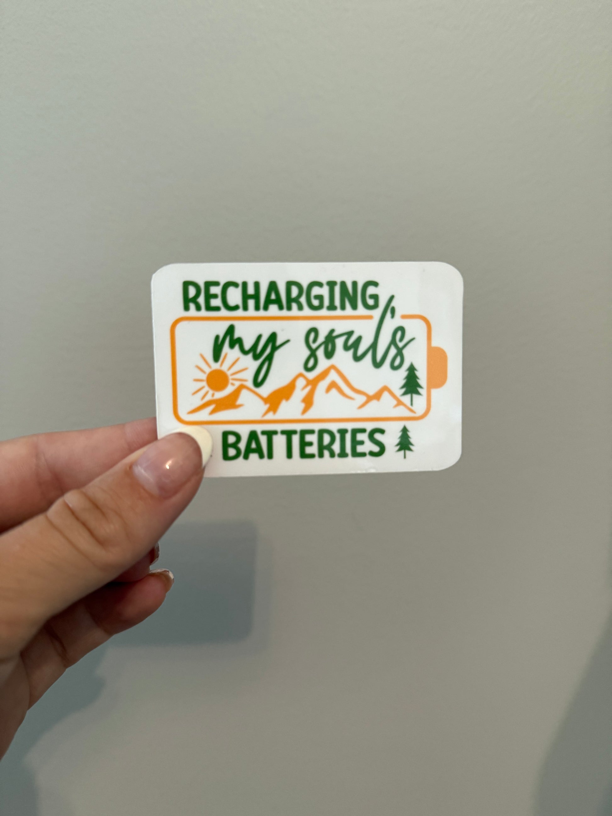 Hand holding a sticker with 'Recharging my souls batteries' text and nature-themed graphics on a plain background.