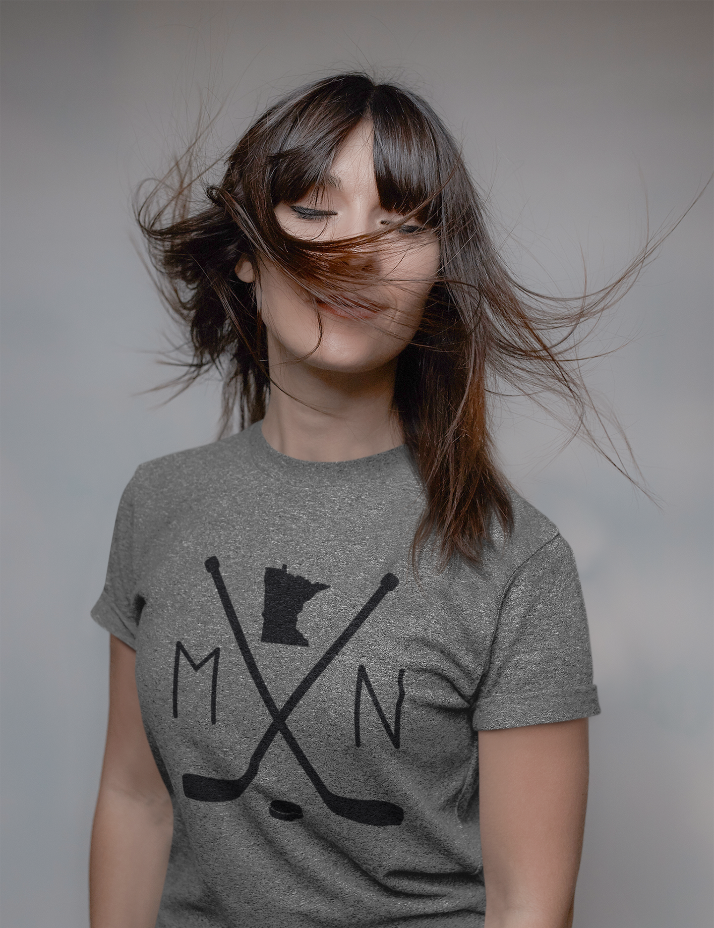 Heather Gray Minnesota Hockey T-Shirt. On the front has a black design with to hockey sticks faced like an X with a puck below in. An M to the left, an N to the right, and an outline of the state of minnesota up top. This is the front view of the shirt on a model whipping her hair around.