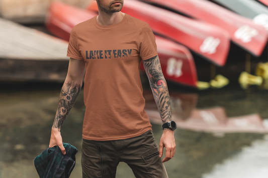 Lake It Easy T-Shirt in orange on a male model in a lake setting with the shirt on, holding a flannel in his hand.