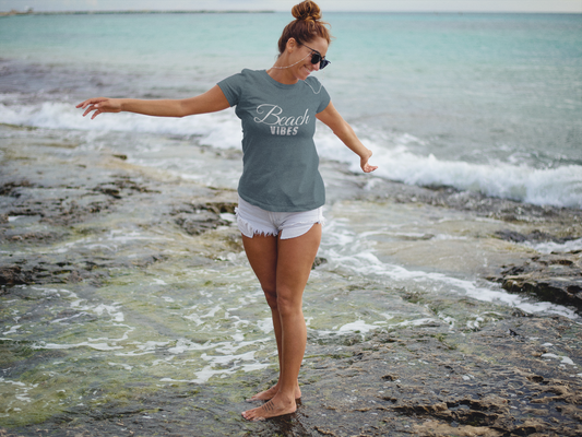 Beach Vibes T-Shirt on a female model oceanside walking through the water. Model has her hair in a bun, sunglasses on with the beach vibes shirt and light jean shorts.