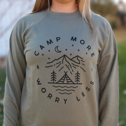 Camp More Worry Less - Pull Over Sweatshirt. Design has "camp more" on top & "worry less" on the bottom curved in a circle with small pine trees separating the words. In the middle includes a tent siting on a bay of water with pine trees standing on both sides of the tent, with mountains in the background and stars aligning with a moon above the mountains. The design is in black on an olive green blank. This is the front view of the sweatshirt on a model (up close view).