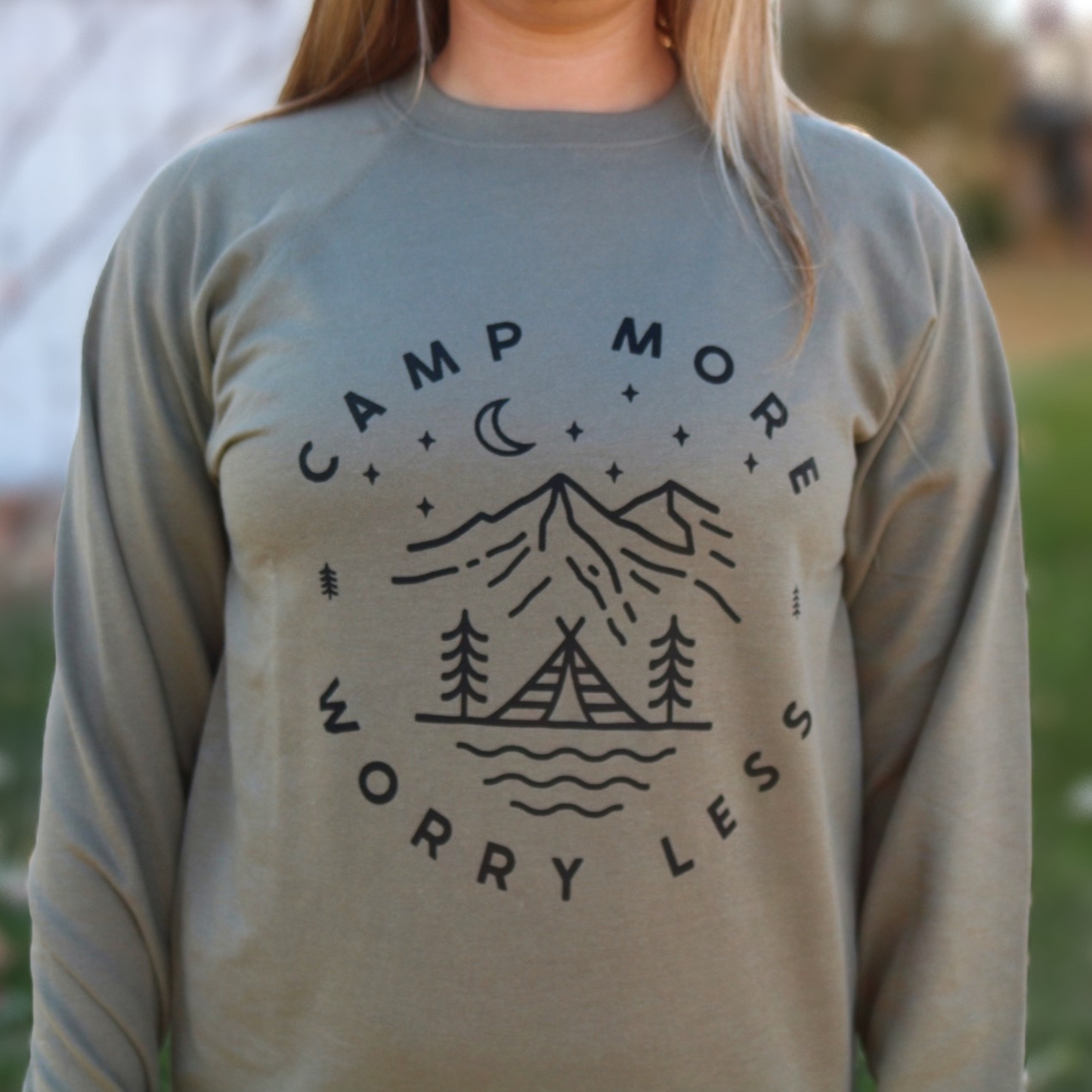 Camp More Worry Less - Pull Over Sweatshirt. Design has "camp more" on top & "worry less" on the bottom curved in a circle with small pine trees separating the words. In the middle includes a tent siting on a bay of water with pine trees standing on both sides of the tent, with mountains in the background and stars aligning with a moon above the mountains. The design is in black on an olive green blank. This is the front view of the sweatshirt on a model (up close view).