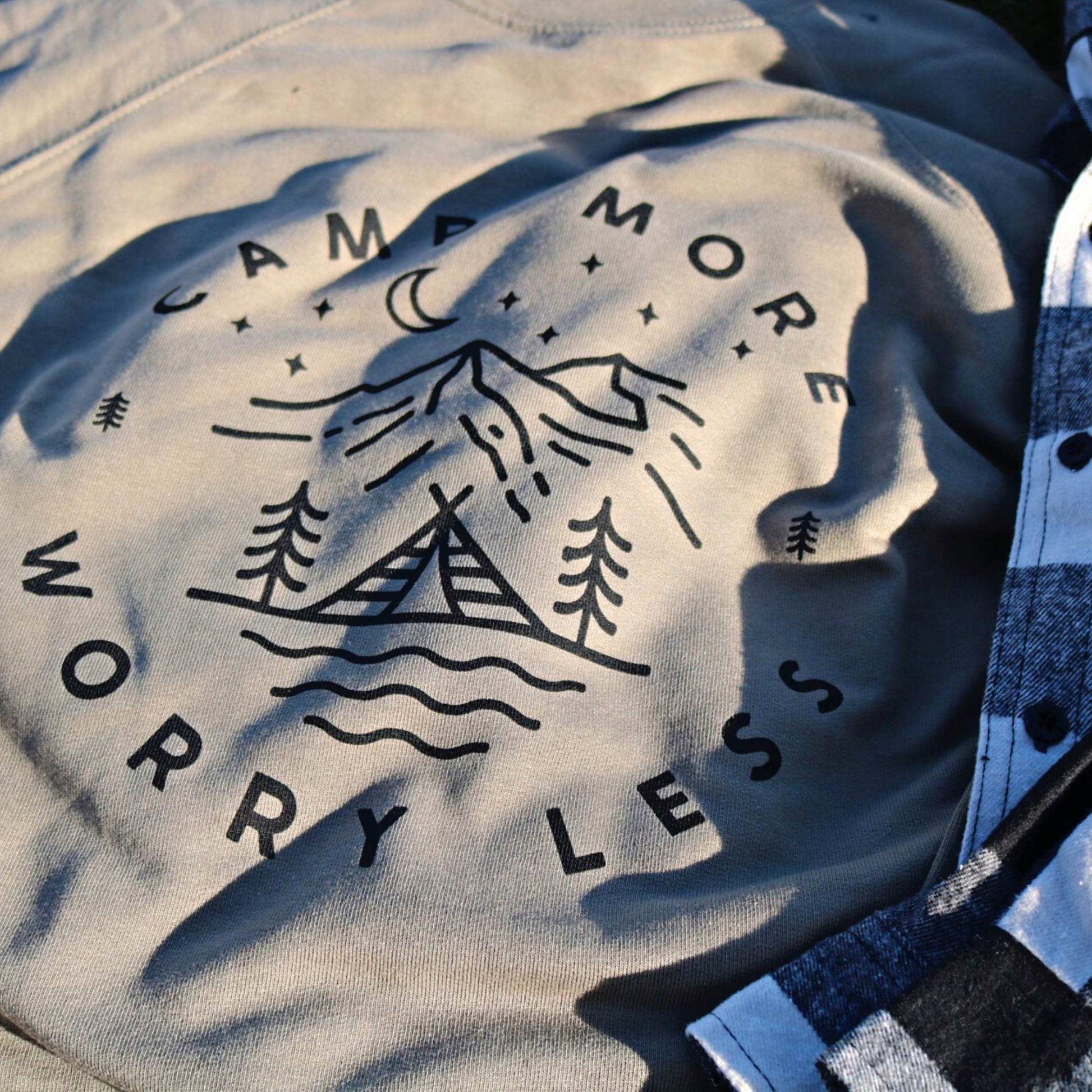 Camp More Worry Less - Pull Over Sweatshirt. Design has "camp more" on top & "worry less" on the bottom curved in a circle with small pine trees separating the words. In the middle includes a tent siting on a bay of water with pine trees standing on both sides of the tent, with mountains in the background and stars aligning with a moon above the mountains. The design is in black on an olive green blank. This is the front and up close view of the sweatshirt, with a black and white flannel next to it.
