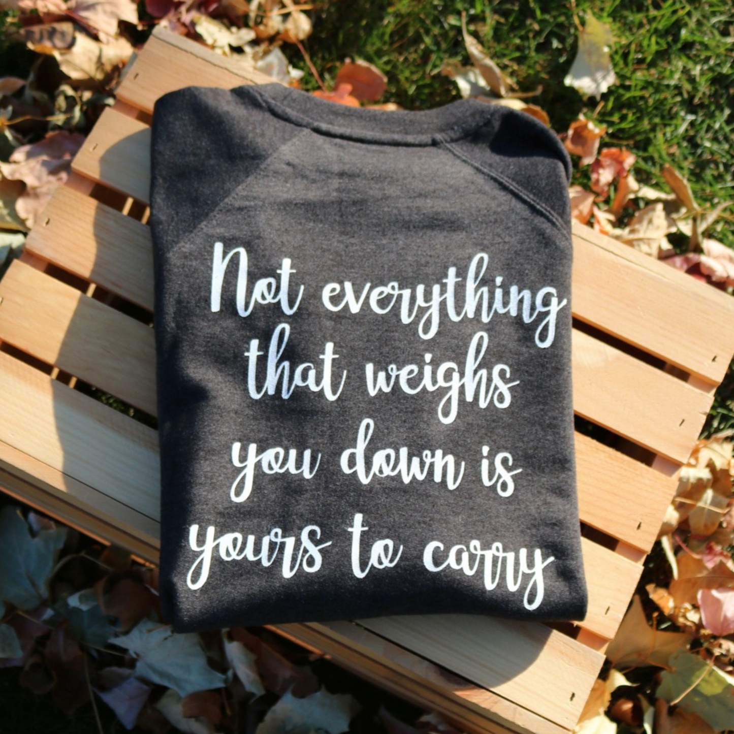 Not Everything That Weighs You Down - Pull Over Sweatshirt in charcoal gray. The back has a white cursive saying of "not everything that weighs you down is yours to carry" with the front corner pocket saying in a cursive font in white of "keep smiling". This is the back view of the sweatshirt, folded on a wooden crate with leaves around it.