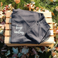 Not Everything That Weighs You Down - Pull Over Sweatshirt in charcoal gray. The back has a white cursive saying of "not everything that weighs you down is yours to carry" with the front corner pocket saying in a cursive font in white of "keep smiling". This is the front view of the sweatshirt folded on a wooden crate with leaves around it.