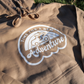 Time For A New Adventure Hooded Sweatshirt. Design is in a cloud bubble with time for a new adventure at the top curved with an off road vehicle in the middle with a rainbow behind it, and adventure below it. This the design in a white on the sandstone tan hoodie. This is a stylized up close shot of the design on the sweatshirt.