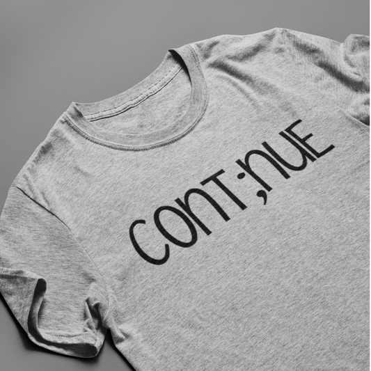 Cont;nue - meaning "my story isn't over". You're the author of your story. Don't end it with a period when you feel stuck. Pause, take it in and continue on. Shirt is in a light gray with black writing. This is the front view of the shirt up close stylized view laid flat on a gray background.