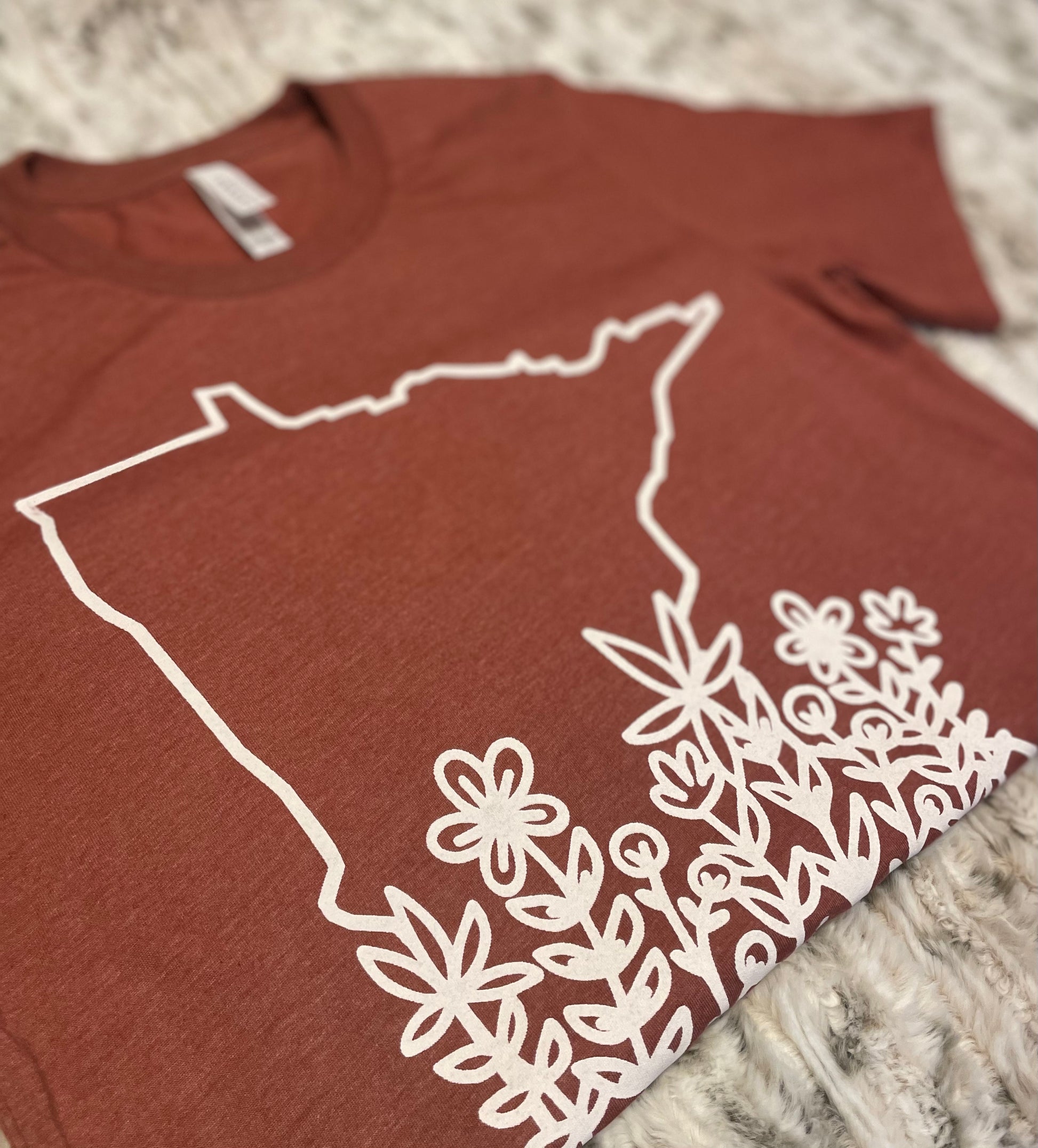 State of Minnesota outline with flowers ranging up the state in white on a heather clay shirt by bella canvas. Front view of the t-shirt (up close to the design)