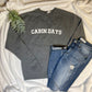 Gray Cabin Days Sweatshirt stylized with destructed denim jeans and a green leafy floral on a cozy white blanket