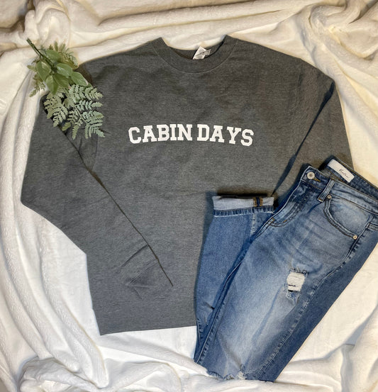 Gray Cabin Days Sweatshirt stylized with destructed denim jeans and a green leafy floral on a cozy white blanket