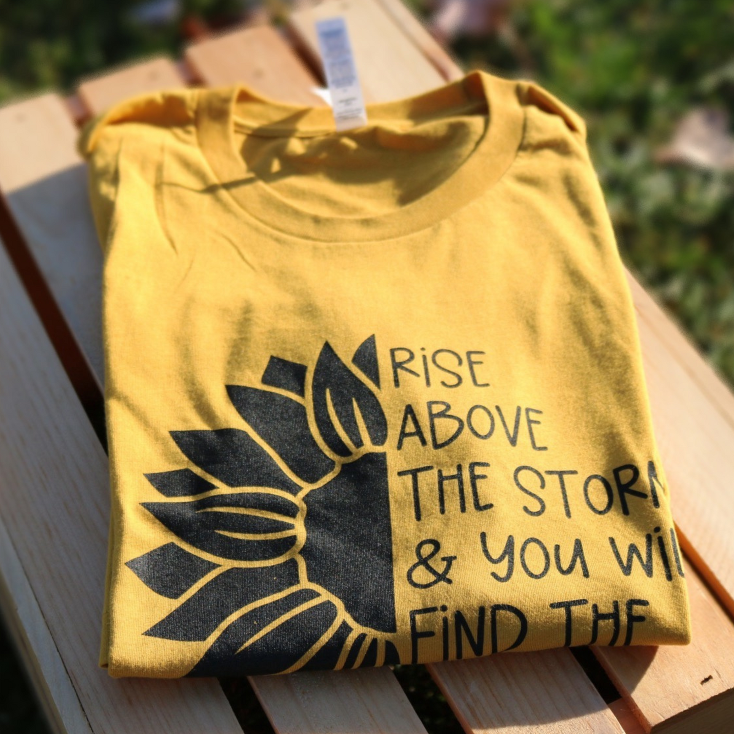 Rise Above The Storm & You Will Find The Sunshine - Yellow T-Shirt - folded front view laying on a wooden crate in a grassy area