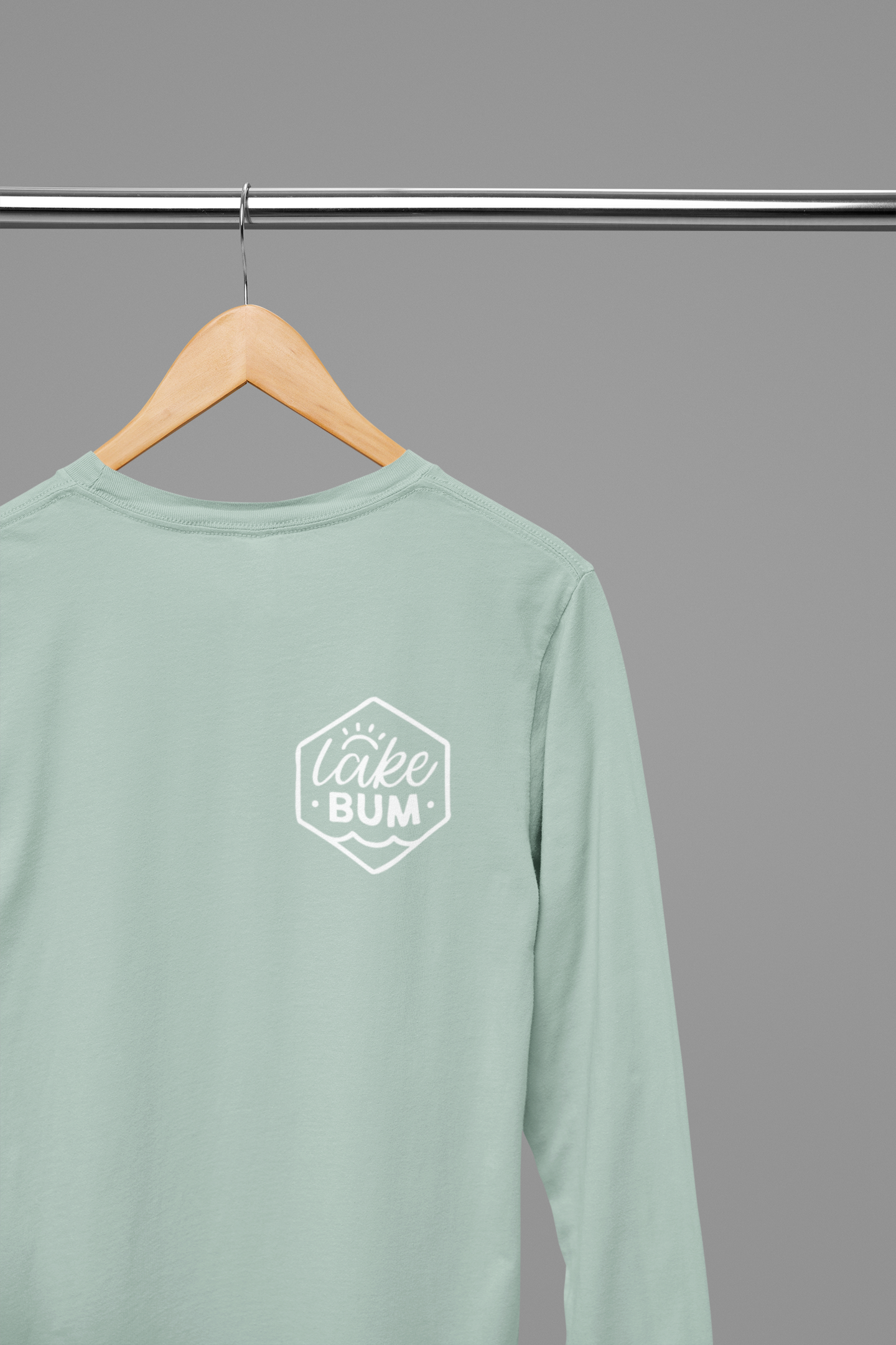 Life is Better at the Lake - Long Sleeve T-Shirt in heather dusty blue. The back of the shirt has a white design with the saying "Life is better at the lake" with lake waves, pine trees, and a loon all squared into a frame. The front has the saying "lake bum" with a lake wave and sun all boxed into a hexagon on the small square pocket (right). This is the front view of the shirt, hanging on a natural wooden hanger hanging on a clothing rack.
