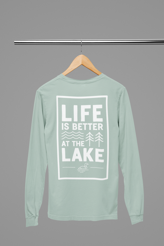 Life is Better at the Lake - Long Sleeve T-Shirt in heather dusty blue. The back of the shirt has a white design with the saying "Life is better at the lake" with lake waves, pine trees, and a loon all squared into a frame. The front has the saying "lake bum" with a lake wave and sun all boxed into a hexagon on the small square pocket (right). This is the back view of the shirt hanging on a natural wooden hanger hanging on a clothing rack.