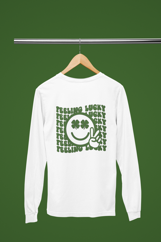 Feeling Lucky Long Sleeve Shirt in white. The back has a repetitive curvy saying "feeling lucky" with a smiley face making a peace sign with the eyes as clovers in green. The front has a four leaf clover pocket sale on the front in green. This is the back view of the shirt hanging on a wooden hanger on a clothes rack with the background green