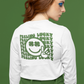 Feeling Lucky Long Sleeve Shirt in white. The back has a repetitive curvy saying "feeling lucky" with a smiley face making a peace sign with the eyes as clovers in green. The front has a four leaf clover pocket sale on the front in green. This is the back view of the shirt on a female model. Shirt is tucked into her green pants standing in front of a green background.