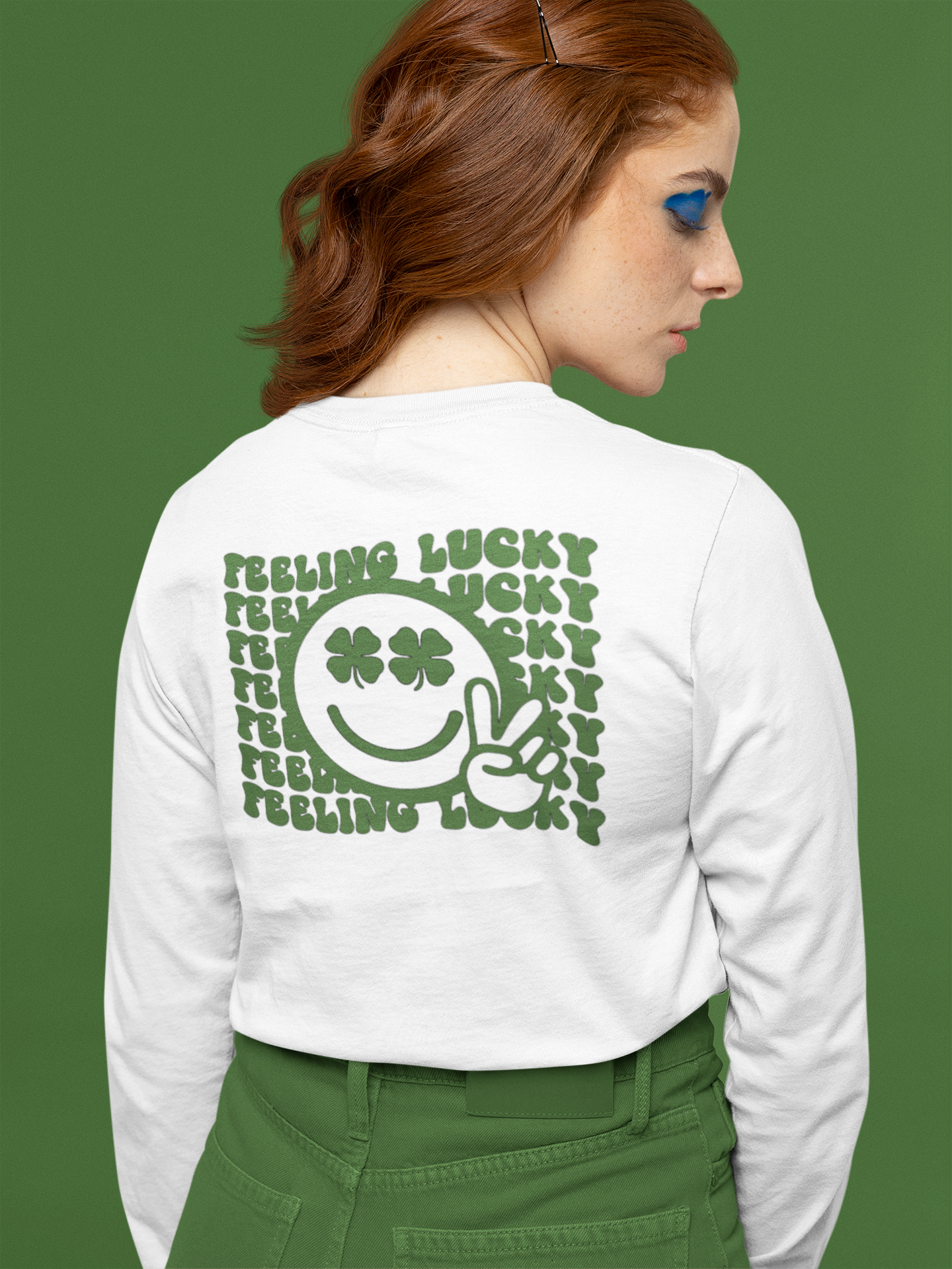 Feeling Lucky Long Sleeve Shirt in white. The back has a repetitive curvy saying "feeling lucky" with a smiley face making a peace sign with the eyes as clovers in green. The front has a four leaf clover pocket sale on the front in green. This is the back view of the shirt on a female model. Shirt is tucked into her green pants standing in front of a green background.