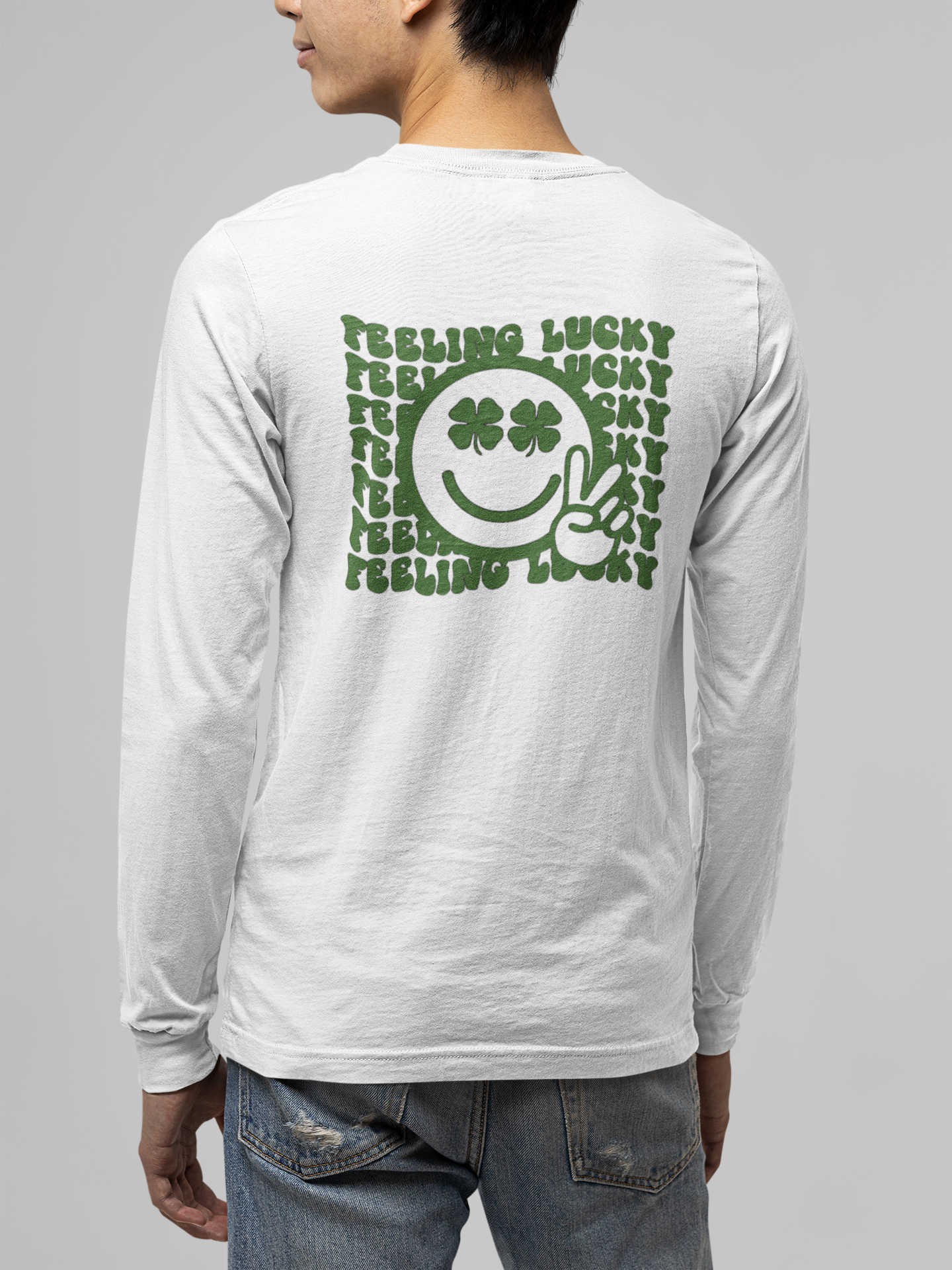Feeling Lucky Long Sleeve Shirt in white. The back has a repetitive curvy saying "feeling lucky" with a smiley face making a peace sign with the eyes as clovers in green. The front has a four leaf clover pocket sale on the front in green. This is the back view of the shirt on a male model.