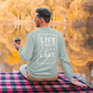Life is Better at the Lake - Long Sleeve T-Shirt in heather dusty blue. The back of the shirt has a white design with the saying "Life is better at the lake" with lake waves, pine trees, and a loon all squared into a frame. The front has the saying "lake bum" with a lake wave and sun all boxed into a hexagon on the small square pocket (right). This is the back view of the shirt on a model holding a cup of coffee at the end of a dock looking at the beautiful fall tree's surrounding the lake.