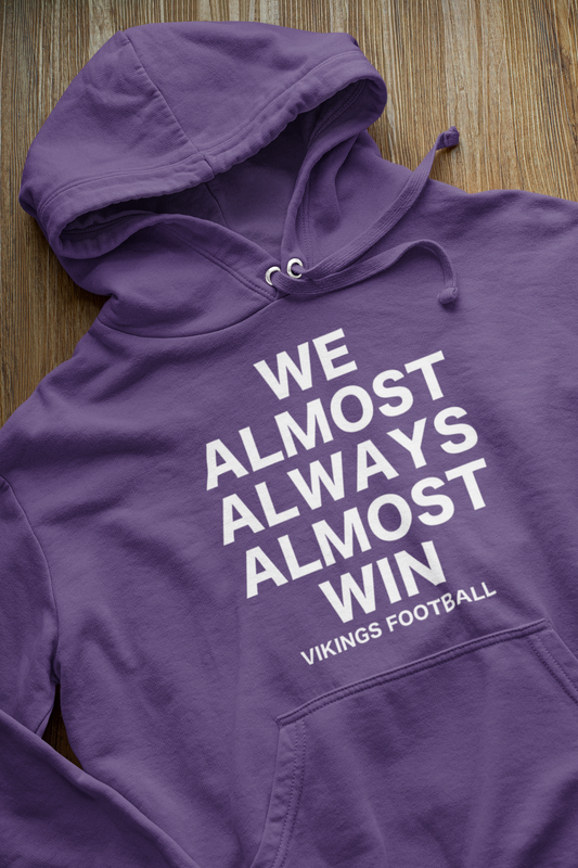 We Almost Always Almost Win Vikings Football Hooded Sweatshirt in purple with white writing. This is the front view of the sweatshirt up close view laying on a hard wood floor.