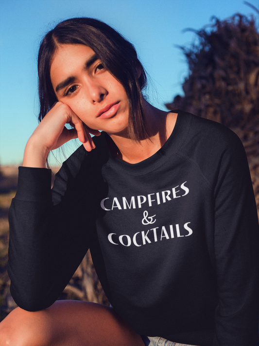 Campfires & Cocktails - Pull Over Sweatshirt. Black unisex crew neck pull over sweatshirt in a lightweight fabric with the saying "campfires & cocktails" in a bold white font. This is the front view of the sweatshirt on a model in an outdoor scene with sun shinning on her face and her hand up on her chin, resting her head on her hand.