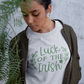 Luck Of The Irish - Pull Over Sweatshirt in a heather light gray. Luck Of The Irish quote on the sweatshirt is a medium green with cute leaves enclosing the design and a small four leaf clover. This is a stylized picture of a women wearing the sweatshirt with a green bomber jacket over the top of it, with some greenery in the front of her.