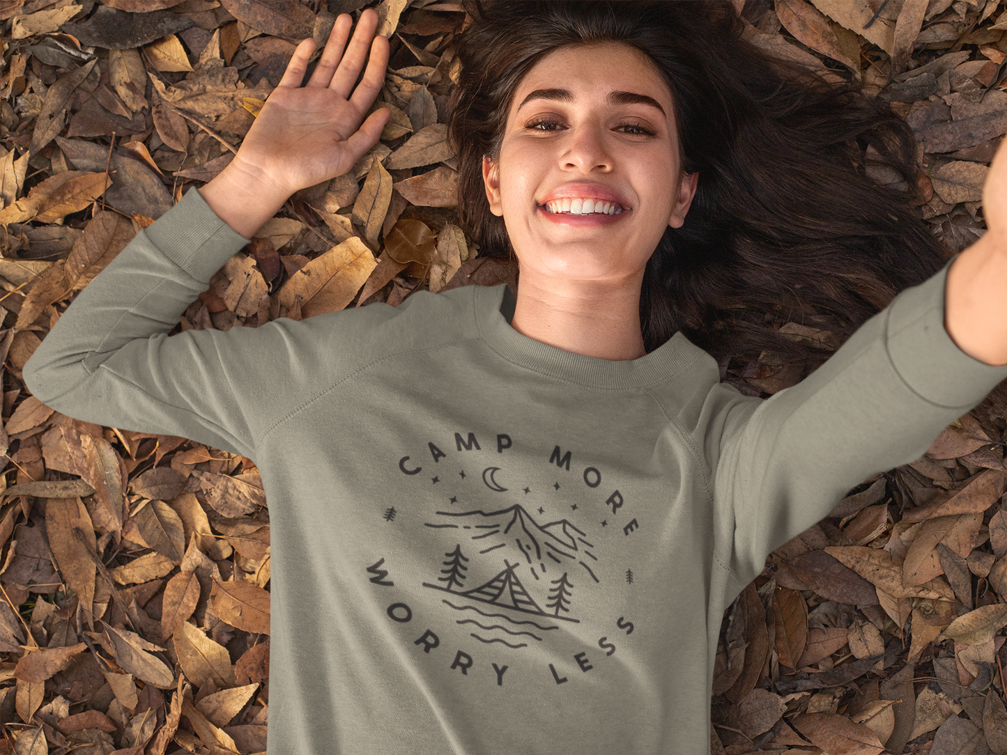 Camp More Worry Less - Pull Over Sweatshirt. Design has "camp more" on top & "worry less" on the bottom curved in a circle with small pine trees separating the words. In the middle includes a tent siting on a bay of water with pine trees standing on both sides of the tent, with mountains in the background and stars aligning with a moon above the mountains. The design is in black on an olive green blank. This is the front view of the sweatshirt on a model laying in fall leaves and smiling.