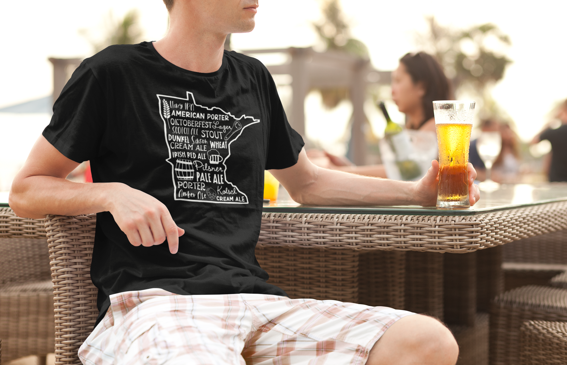 Male model sitting at a table with a beer in his hand and wearing the black Minnesota Beers - T-Shirt with hazy ipa, American porter, Oktoberfest, lager, scotch ale, stout, dunkel, saison, wheat, cream ale, Irish red ale, pilsner, pale ale, porter, kolsch, amber ale, cream ale on the front in the state of Minnesota with icons of a hop, glass of beer, barrel of beer, bottle opener, and a wheat strand.