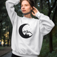 Moonlight Mountain - Pull Over Sweatshirt. The design is in black with a half moon, pine trees mountains and stars sitting inside the moon. This design is on a white sweatshirt. The image is of a girl with it on holding her hair up and posing in the middle of a walking trail with trees surrounding her.