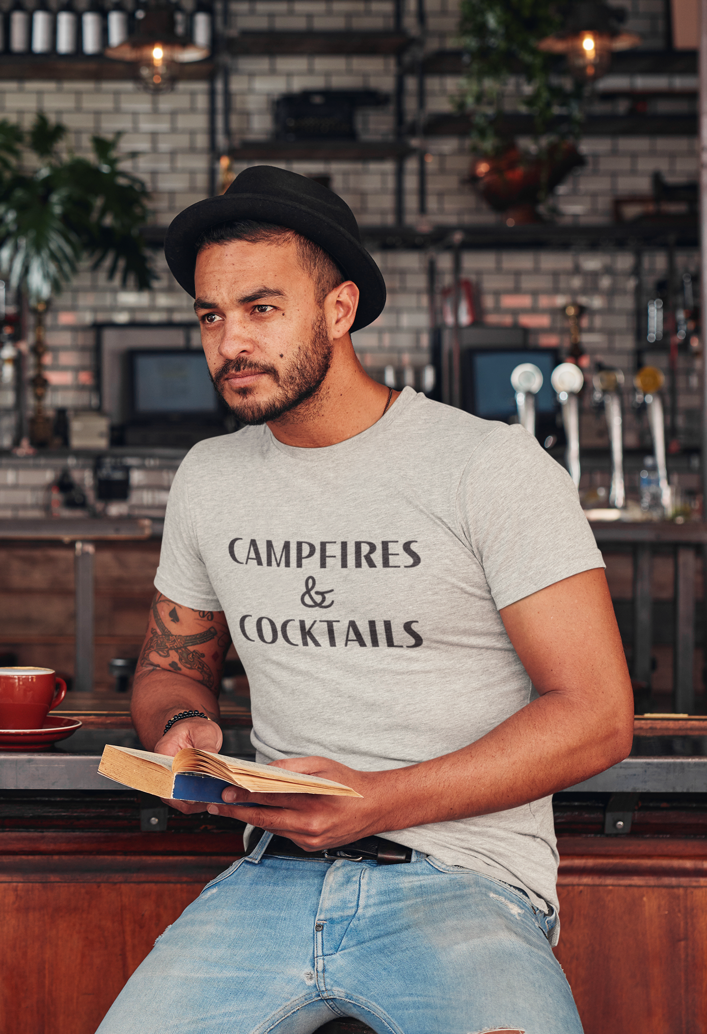 Campfires & Cocktails - T-Shirt in Heather oatmeal. The front of the shirt has a black bold font with campfires & cocktails on it. This is the front view of the shirt on a male model holding a book in a bar like setting with black lid hat and light dem jeans.