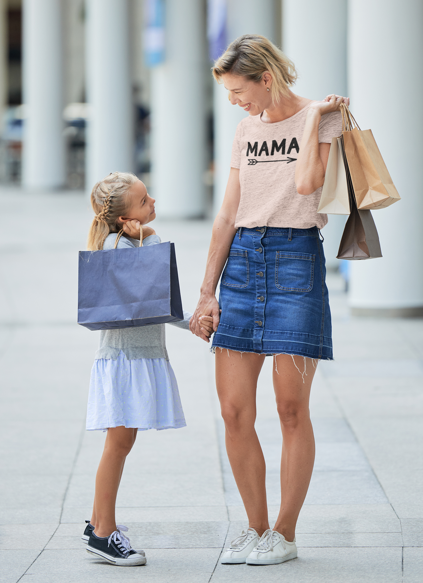 Mama - T-Shirt in heather prism peach. Mama is in a black design with an arrow below it pointing to the right. This is an image of a mom and her daughter shopping with her holding shopping bags. Mom has the Mama shirt on with denim jean skirt and tennis, with the little girl holding her hand wearing a light purple dress, gray sweater and black converse tennis shoes.