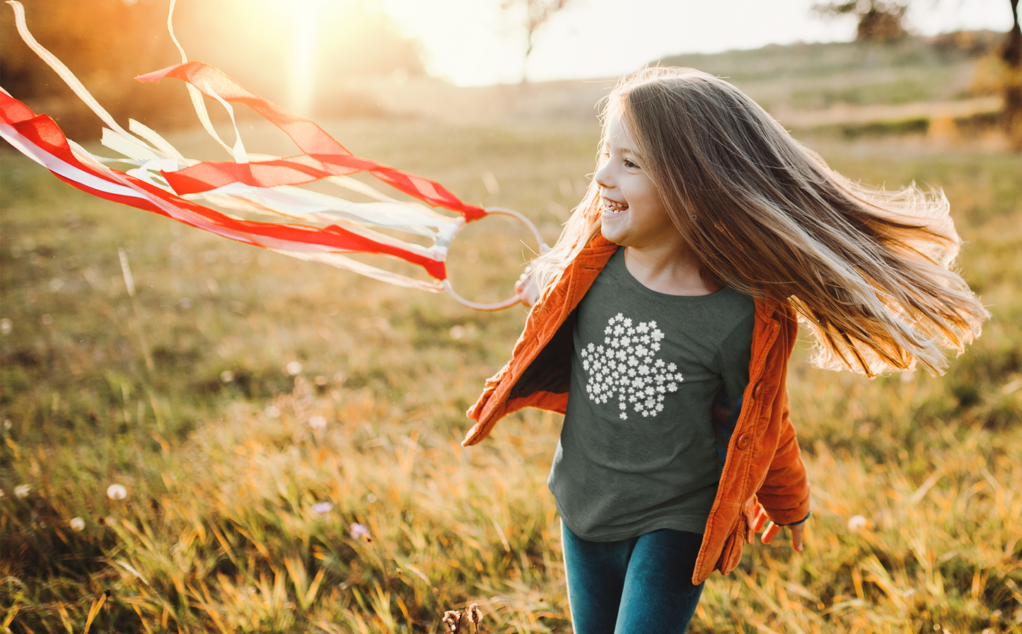 Four-Leaf Clover T-Shirt in heather forest. The front of the t-shirt has a large four leaf clover with mini four leaf clovers making the shape of the larger clover in white. This is the front view of the shirt on a little girl with with an orange coat on over it running through a grassy field with a kite like item in her hand.