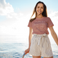 Life Is Better By The Lake - T-Shirt in Mauve with white writing on a model with cute cream and black striped boat shorts. Model is standing on the edge of the boat hanging onto the ladder with lake & beautiful blue sky above her. Model is smiling with her brown hair flowing in the wind.