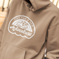 Time For A New Adventure Hooded Sweatshirt. Design is in a cloud bubble with time for a new adventure at the top curved with an off road vehicle in the middle with a rainbow behind it, and adventure below it. This the design in a white on the sandstone tan hoodie. This is a stylized up close shot of the design on the sweatshirt on a model looking opposite from the camera.