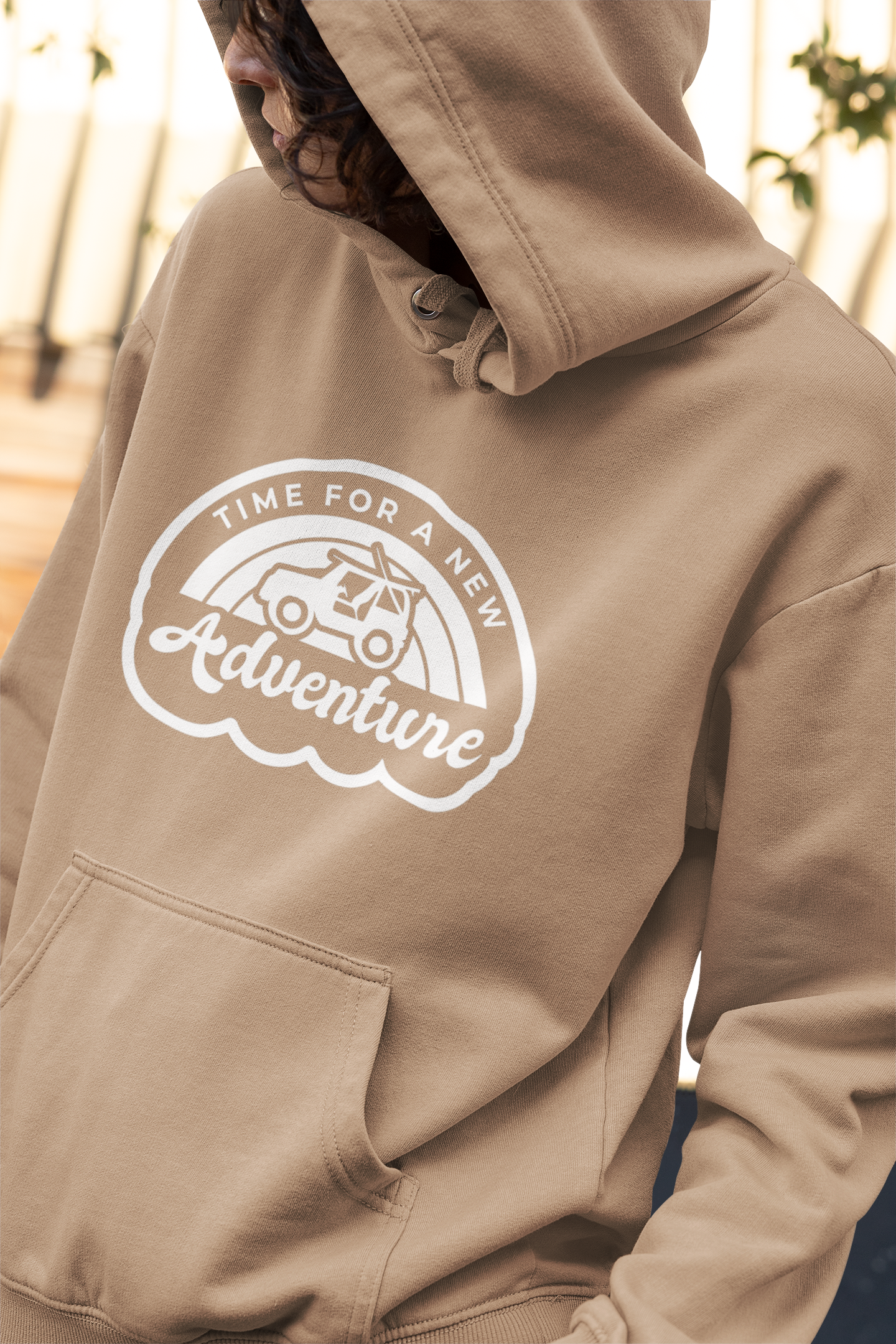 Time For A New Adventure Hooded Sweatshirt. Design is in a cloud bubble with time for a new adventure at the top curved with an off road vehicle in the middle with a rainbow behind it, and adventure below it. This the design in a white on the sandstone tan hoodie. This is a stylized up close shot of the design on the sweatshirt on a model looking opposite from the camera.