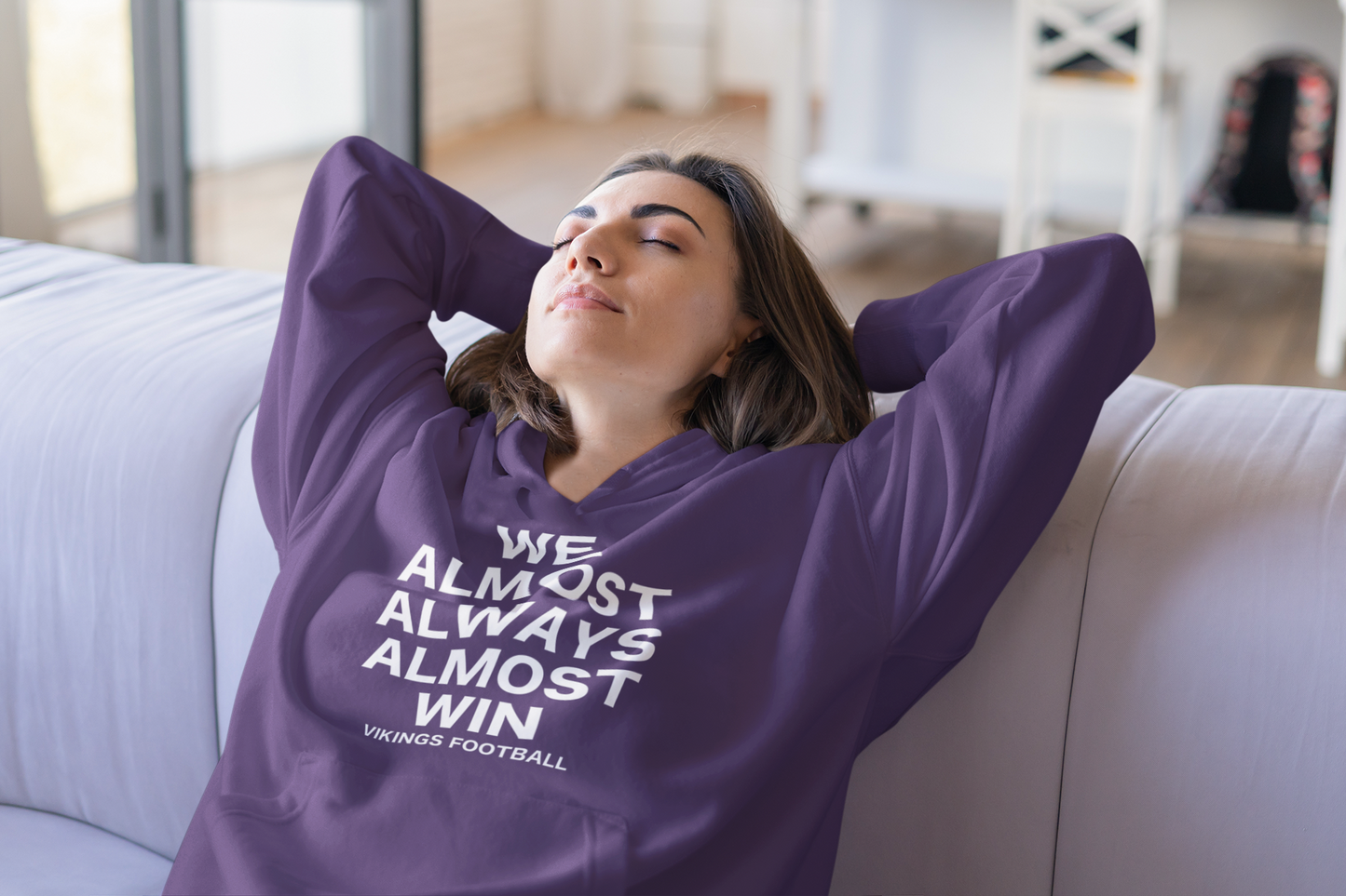 We Almost Always Almost Win Vikings Football Hooded Sweatshirt in purple with white writing. This is the front view of the sweatshirt on a model. She is sitting on a couch with her hands and arms back behind her head, closing her eyes in disappointment.
