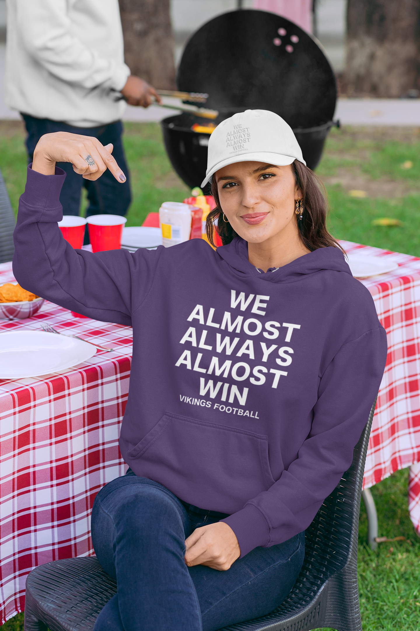We Almost Always Almost Win Vikings Football Hooded Sweatshirt in purple with white writing. This is the front view of the sweatshirt on a woman model sitting in a backyard bbq scene pointing to her sweatshirt with a white hat on.