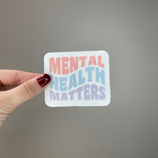 The Mental Health Matters Sticker featuring a curvy light pink, blue and purple bold font on a gray background with someone holding the sticker.