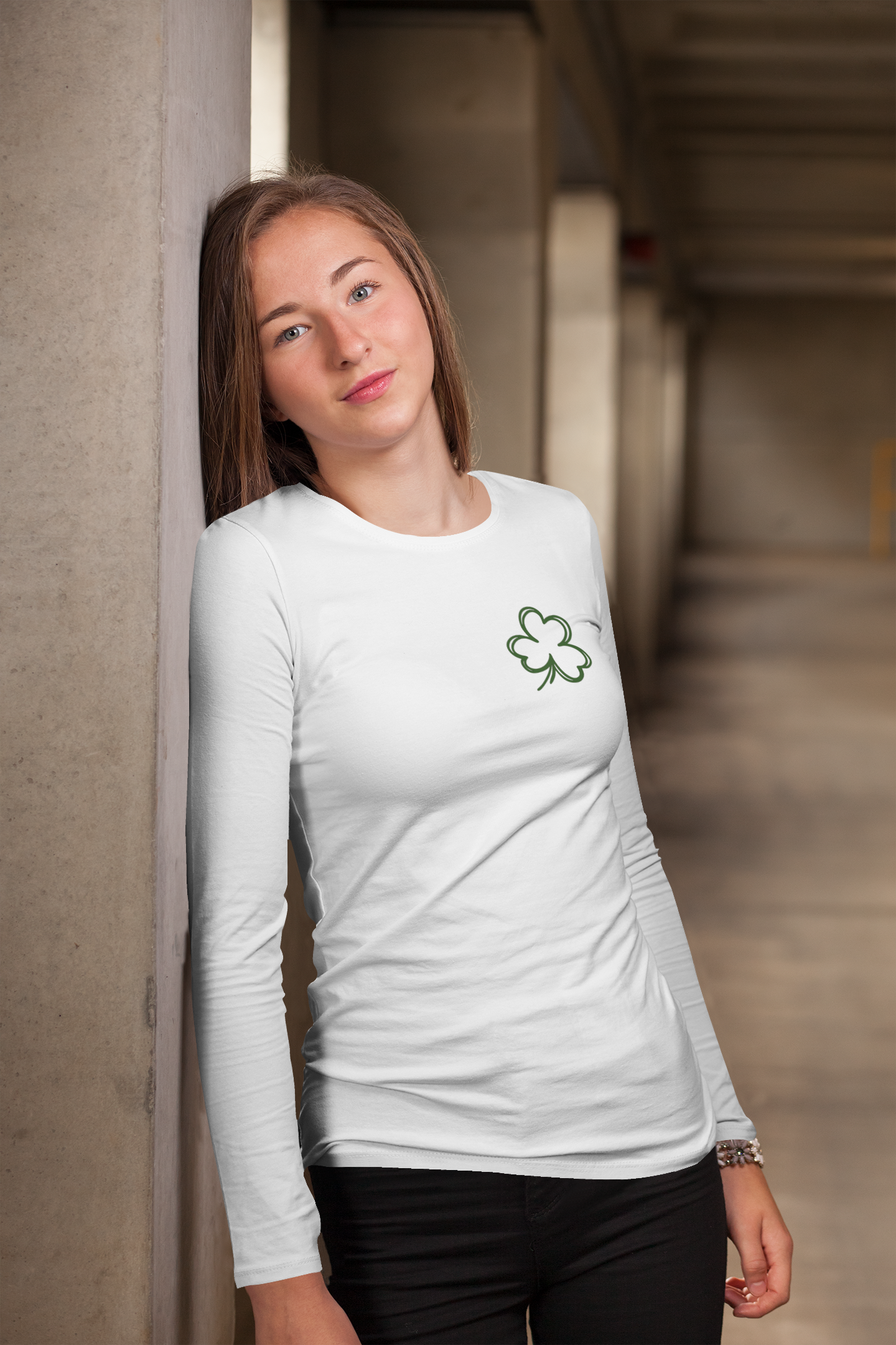 Feeling Lucky Long Sleeve Shirt in white. The back has a repetitive curvy saying "feeling lucky" with a smiley face making a peace sign with the eyes as clovers in green. The front has a four leaf clover pocket sale on the front in green. This is the front view of the shirt on a female model leaning up against a cement pillar.