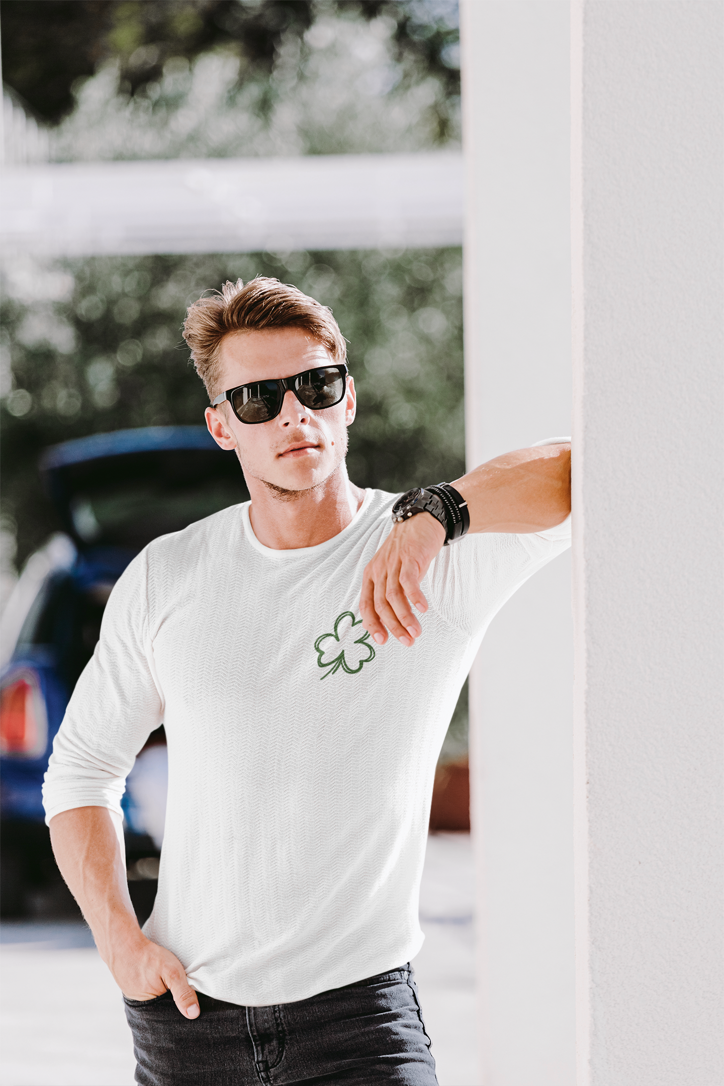 Feeling Lucky Long Sleeve Shirt in white. The back has a repetitive curvy saying "feeling lucky" with a smiley face making a peace sign with the eyes as clovers in green. The front has a four leaf clover pocket sale on the front in green. This is the front view of the shirt on a male model with sunglasses on leaning up against a pillar outside.