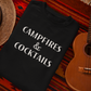 Campfires & Cocktails - Pull Over Sweatshirt. Black unisex crew neck pull over sweatshirt in a lightweight fabric with the saying "campfires & cocktails" in a bold white font. This is the front view of the sweatshirt folded on a orange dark based aztec blanket underneath it, a straw hat to the left of the sweatshirt and a mini ukulele to the right of the sweatshirt.