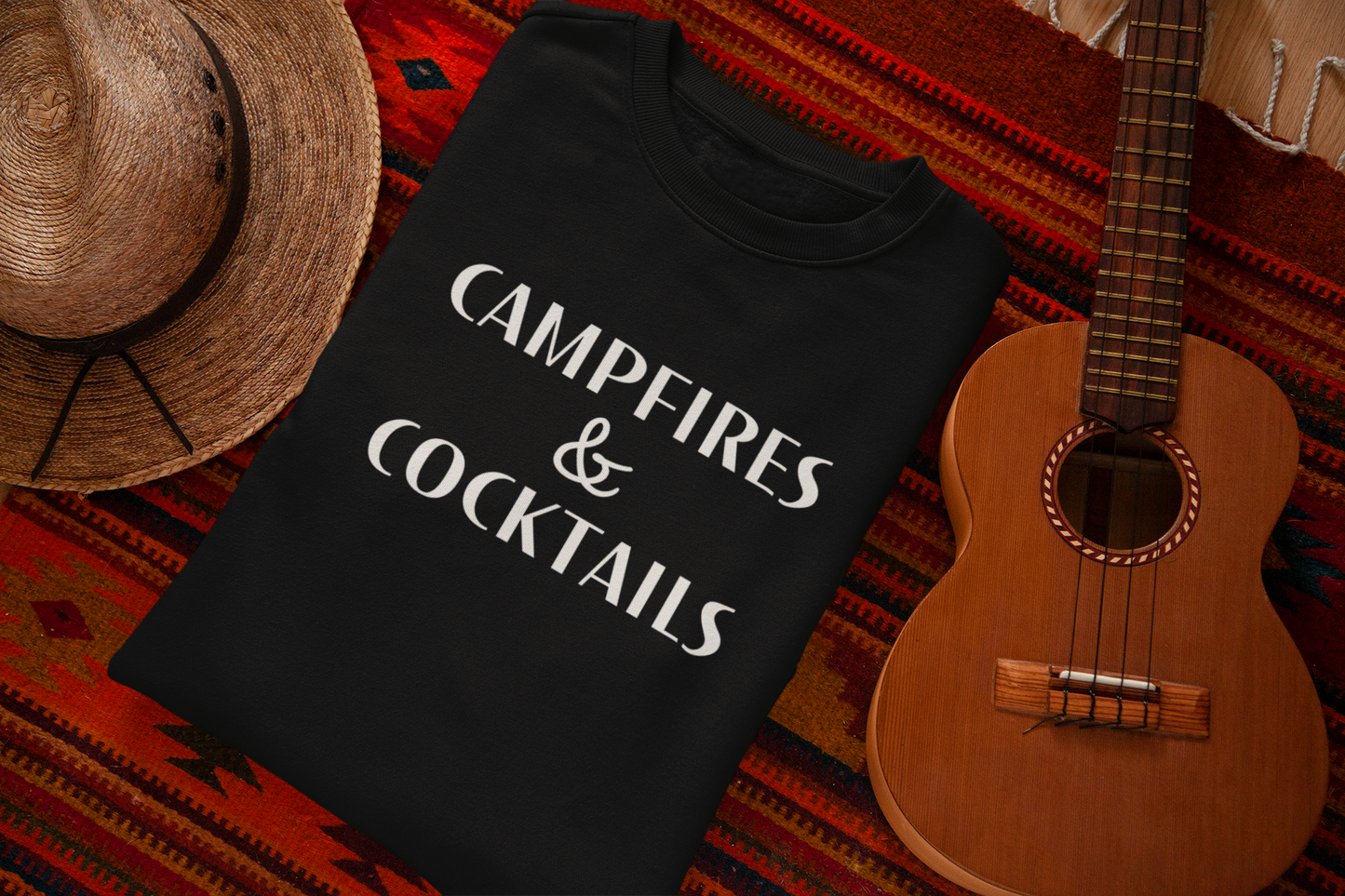 Campfires & Cocktails - Pull Over Sweatshirt. Black unisex crew neck pull over sweatshirt in a lightweight fabric with the saying "campfires & cocktails" in a bold white font. This is the front view of the sweatshirt folded on a orange dark based aztec blanket underneath it, a straw hat to the left of the sweatshirt and a mini ukulele to the right of the sweatshirt.