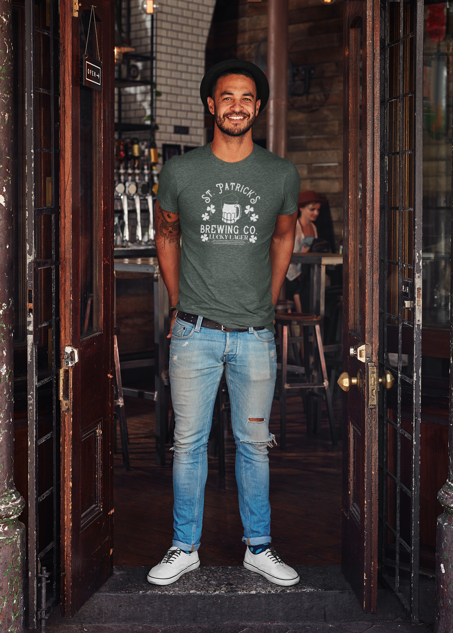 St. Patricks Brewing Co. T-Shirt in heather forest. The front has a white design with the saying "St. Patrick's Brewing Co. Lucky Lager" with a glass of beer and four leaf clovers surrounding the saying. This is the front view of the shirt on a male model standing between brown wooden doors of bar or pub smiling at the camera with his hands in his back pockets.