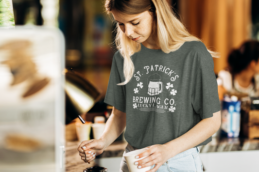 St. Patricks Brewing Co. T-Shirt in heather forest. The front has a white design with the saying "St. Patrick's Brewing Co. Lucky Lager" with a glass of beer and four leaf clovers surrounding the saying. This is the front view of the shirt on a female model with a cup in her hand and spoon in the other.