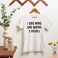 I Like Wine & Maybe 3 People - White T-Shirt in black writing. This is an image of the t-shirt hanging on a brown wooden hanger hanging on a brown wooden hanger rack. To the left of it sits a brown table with modular cream shaped vases and green eucalyptus greenery inside it and white candle. To the right is a pink circular vase with yellow buds coming out of it. All on a creamy background.