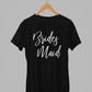 Bridesmaid T-Shirt in black with a white script font writing on the front of the tee. This is the front view of the t-shirt hanging on a wooden hanger hanging on a silver metal clothing rack