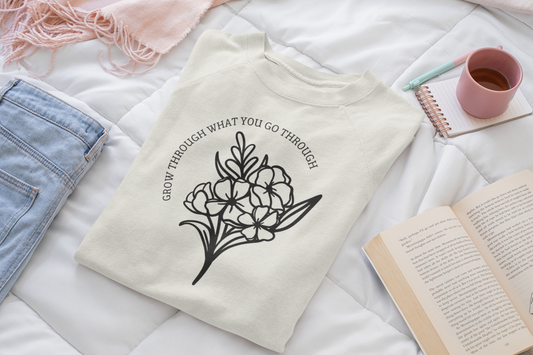 Grow Through What You Go Through - Pull Over Sweatshirt. Sweatshirt is in cream and on the front it has the saying of "grow through what you go through" curved around a bouquet of flowers below it in black. This is the front view of the sweatshirt folded on a white quilted blanket with a pink scarf, light denim jeans on the left, a notebook, pen, cup of coffee and book on the right.