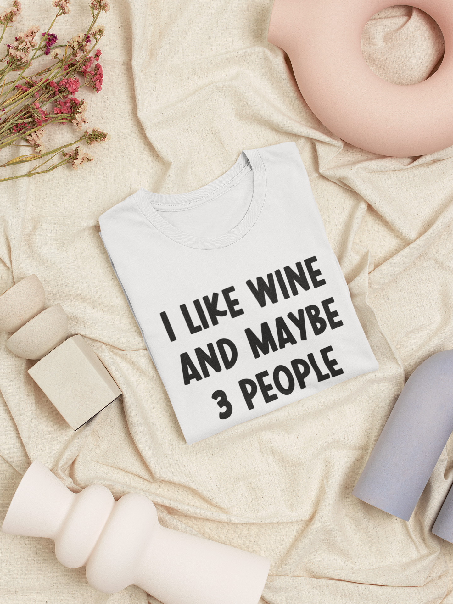 I Like Wine & Maybe 3 People - White T-Shirt in black writing. This is an image of the t-shirt folded in a square on a cream blanket, with a stringy pink flower and cream, blush pink, and light blue objects next to it.